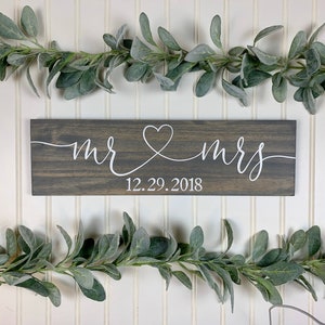 May include: A rectangular wooden sign with the words "mr & mrs" in white script, connected by a heart symbol. The date "12.29.2018" is below. The sign is dark grey and is surrounded by green leafy garlands.