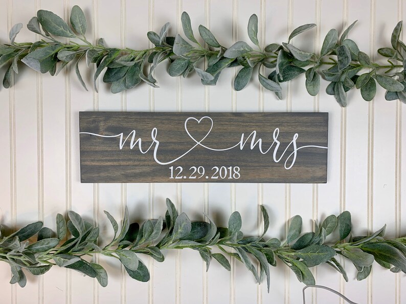 May include: A rectangular wooden sign with the words "mr & mrs" in white script, connected by a heart symbol. The date "12.29.2018" is below. The sign is dark grey and is surrounded by green leafy garlands.