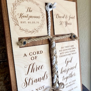 May include: Wooden cross-shaped wall art with a dark brown frame. The light-colored wood features engraved text and a rope cross design. Text includes names, dates, and Bible verses. A unique wedding or anniversary gift.