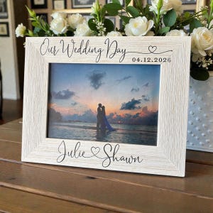 May include: A rectangular white wood-grain picture frame featuring the black text Our Wedding Day, the date 04.12.2026, and the names Julie and Shawn with heart accents. The frame holds a photograph of a couple standing on a beach at sunset.