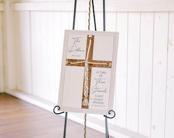 May include: A white sign with a brown wooden cross and a braided rope. The sign reads "The Robbins EST. 2023" and "A cord of three strands is not easily broken. Ecclesiastes 4:12".