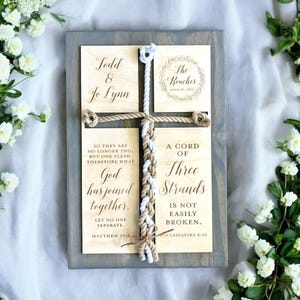 May include: A wooden sign with a rope braid in the center. The sign has four plaques with the names "Todd & Jo Lynn" and "The Rouches" along with the date "June 18, 2014". The plaques also have the text "A Cord of Three Strands is Not Easily Broken" and a Bible verse from Matthew 19:6 and Ecclesiastes 4:12.