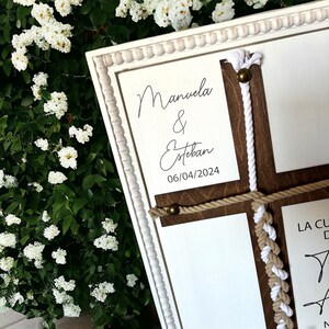 May include: White wooden frame with a brown wooden cross inside. The cross is decorated with white and brown rope. The frame has a white card with the text "Manuela & Esteban 06/04/2024" on it. The frame is a wedding sign.
