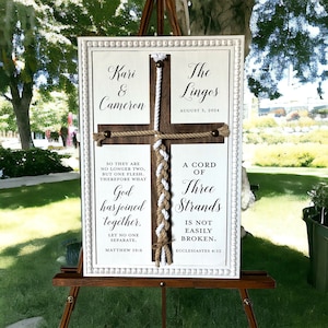 May include: A white sign with a wooden cross and a braided rope design. The sign reads "Kari & Cameron" and "The Lingos" with the date "August 3, 2024". The sign also includes the Bible verses "So they are no longer two, but one flesh. Therefore what God has joined together, let no one separate. Matthew 19:6" and "A cord of three strands is not easily broken. Ecclesiastes 4:12".