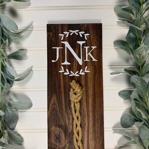May include: A wooden sign with the initials "JNK" in white lettering surrounded by a wreath of leaves. A brown rope hangs from the sign.