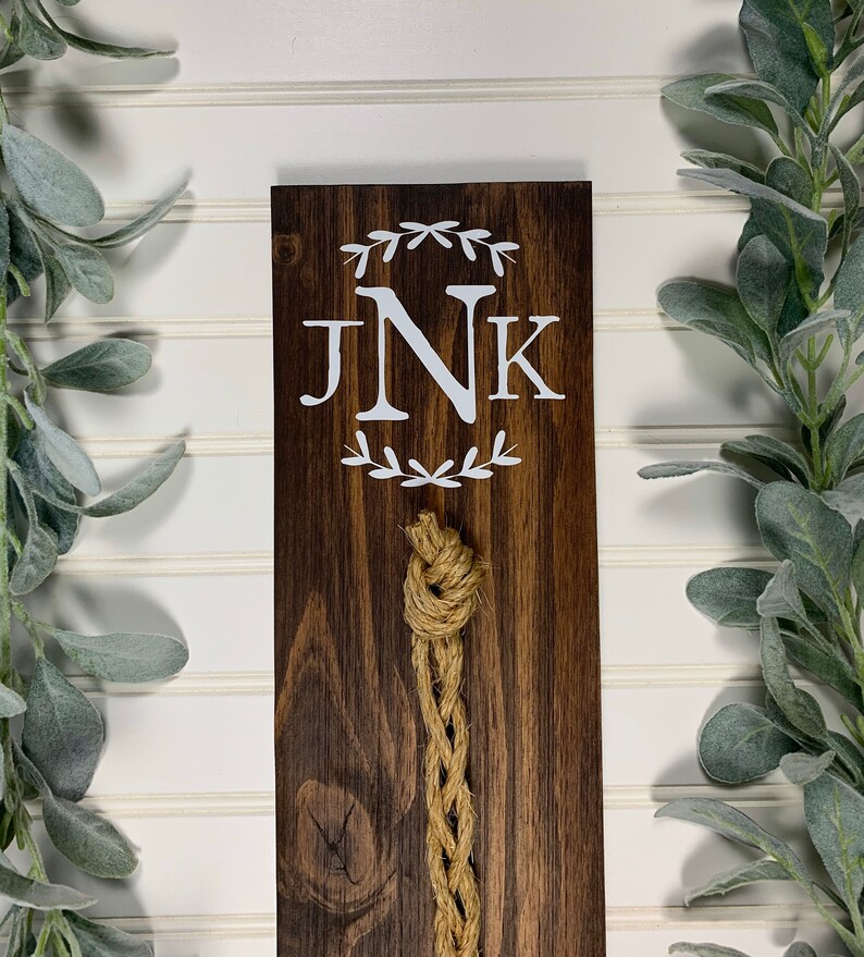 May include: A wooden sign with the initials "JNK" in white lettering surrounded by a wreath of leaves. A brown rope hangs from the sign.