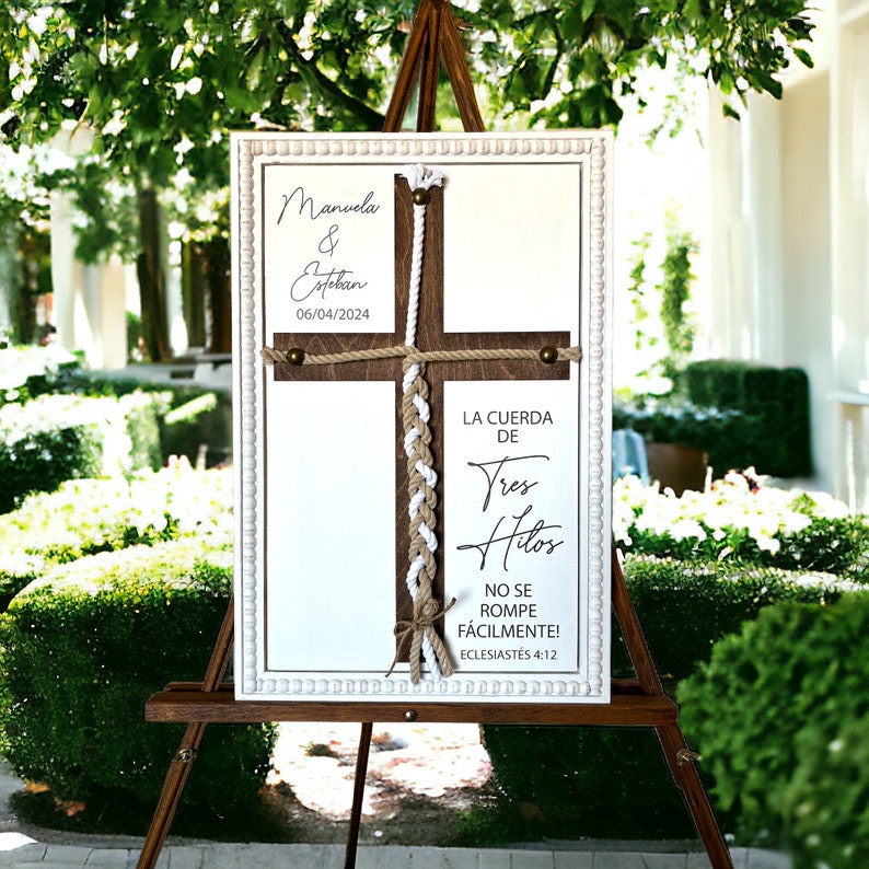 May include: A white wooden sign with a brown wooden cross in the centre. The sign has a white border with a beaded design. The cross is decorated with a white and brown rope. The sign reads "Manuela & Esteban 06/04/2024" and "LA CUERDA DE Tres Hilos NO SE ROMPE F&Aacute;CILMENTE! ECLESIAST&Eacute;S 4:12".