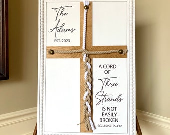 May include: White wooden sign with a white rope and a brown rope intertwined in the shape of a cross. The sign reads "The Adams EST. 2023" and "A cord of three strands is not easily broken. Ecclesiastes 4:12".