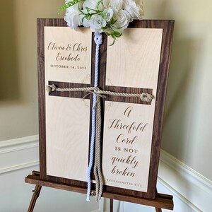 May include: A wooden sign with a cross design and two ropes tied in a knot. The sign is engraved with the text "Olivia & Chris Escobedo, October 14, 2023, A Threefold Cord Is Not Quickly Broken, Ecclesiastes 4:12".