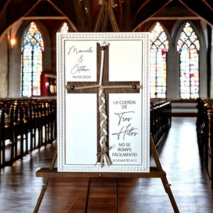 May include: A white sign with a wooden cross and a braided rope. The sign reads "Manuela & Estekan 06/04/2024" and "La Cuerda de Tres Hilos No Se Rompe F&aacute;cilmente! Ecclesiastes 4:12".