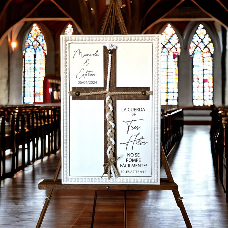 May include: A white sign with a wooden cross and a braided rope. The sign reads "Manuela & Estekan 06/04/2024" and "La Cuerda de Tres Hilos No Se Rompe F&aacute;cilmente! Ecclesiastes 4:12".
