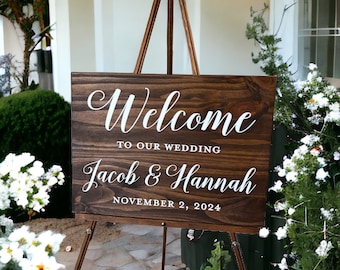 May include: A wooden welcome sign with white text that reads "Welcome to our wedding Jacob & Hannah November 2, 2024". The sign is on a wooden easel.