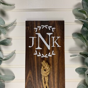 May include: A dark brown wooden sign with the initials "JNK" in white lettering surrounded by a wreath of leaves. A knotted rope hangs from the bottom of the sign.
