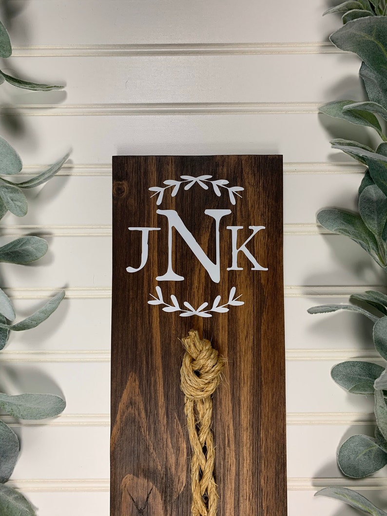 May include: A dark brown wooden sign with the initials "JNK" in white lettering surrounded by a wreath of leaves. A knotted rope hangs from the bottom of the sign.