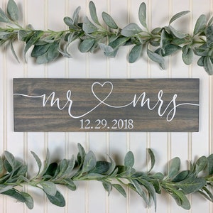 May include: A grey wooden sign with white script reading "mr & mrs", joined by a heart. The date "12.29.2018" is below. The sign is decorated with faux greenery, and is a wedding or anniversary decoration.