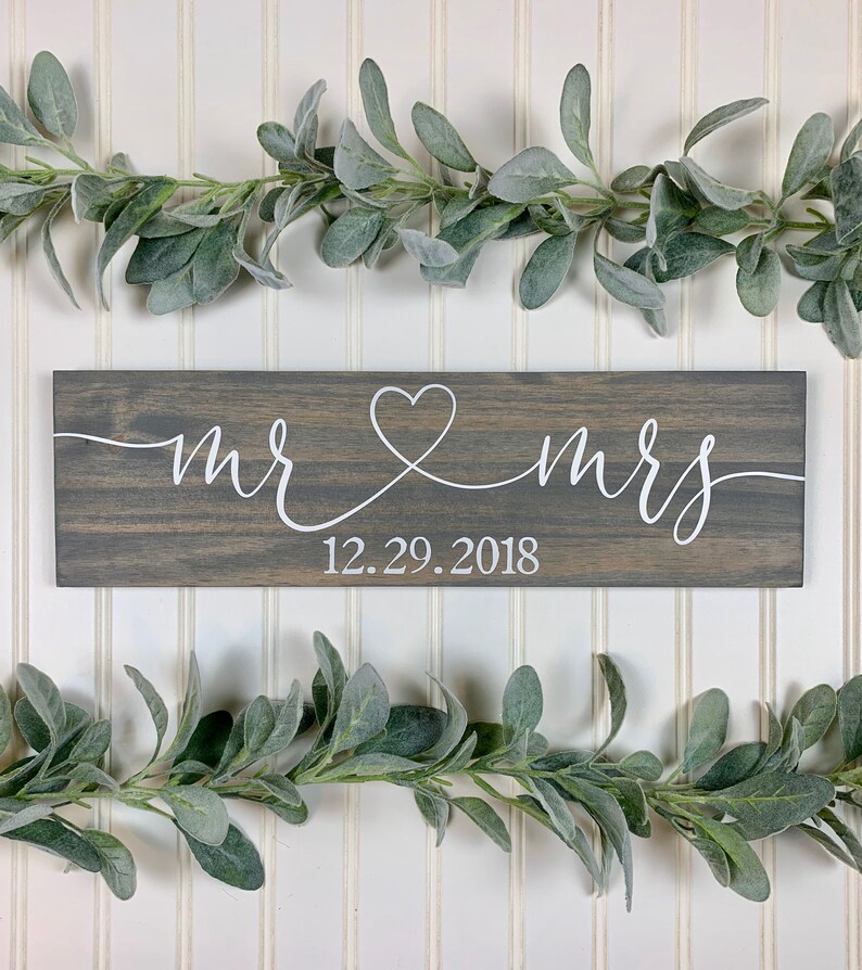 May include: A grey wooden sign with white script reading "mr & mrs", joined by a heart. The date "12.29.2018" is below. The sign is decorated with faux greenery, and is a wedding or anniversary decoration.