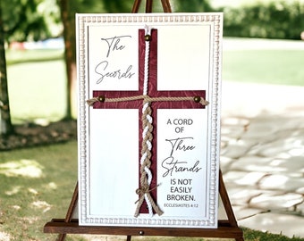 May include: A white wooden sign with a red cross and a braided rope design. The sign reads "The Secords" and "A cord of three strands is not easily broken. Ecclesiastes 4:12".