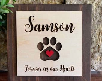 May include: A wooden sign with the name "Samson" in script above a paw print with a red heart. The text "Forever in our Hearts" is below the paw print. The sign has a light wood center and a dark brown frame.