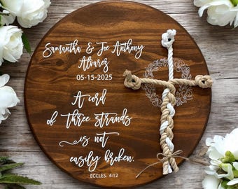 May include: A round, wooden wedding sign with a dark brown stain. The sign features white text with the names "Samantha & Joe Anthony Alvarez" and the date "05-15-2025". A rope cross is attached to the sign, with the quote "A cord of three strands is not easily broken."