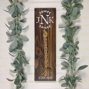 May include: A wooden sign with a white monogram "JNK" and a date "2/20/2020". A brown rope is tied in a knot on the sign.