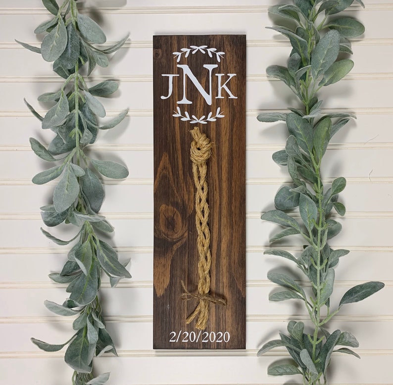 May include: A wooden sign with a white monogram "JNK" and a date "2/20/2020". A brown rope is tied in a knot on the sign.