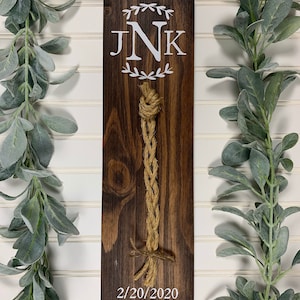 May include: A wooden sign with the initials "JNK" in a white wreath, a braided rope hanging from the top, and the date "2/20/2020" written below.