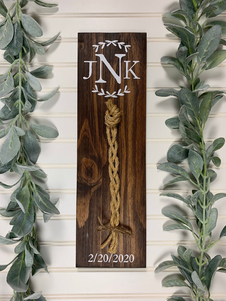 May include: A wooden sign with the initials "JNK" in a white wreath, a braided rope hanging from the top, and the date "2/20/2020" written below.