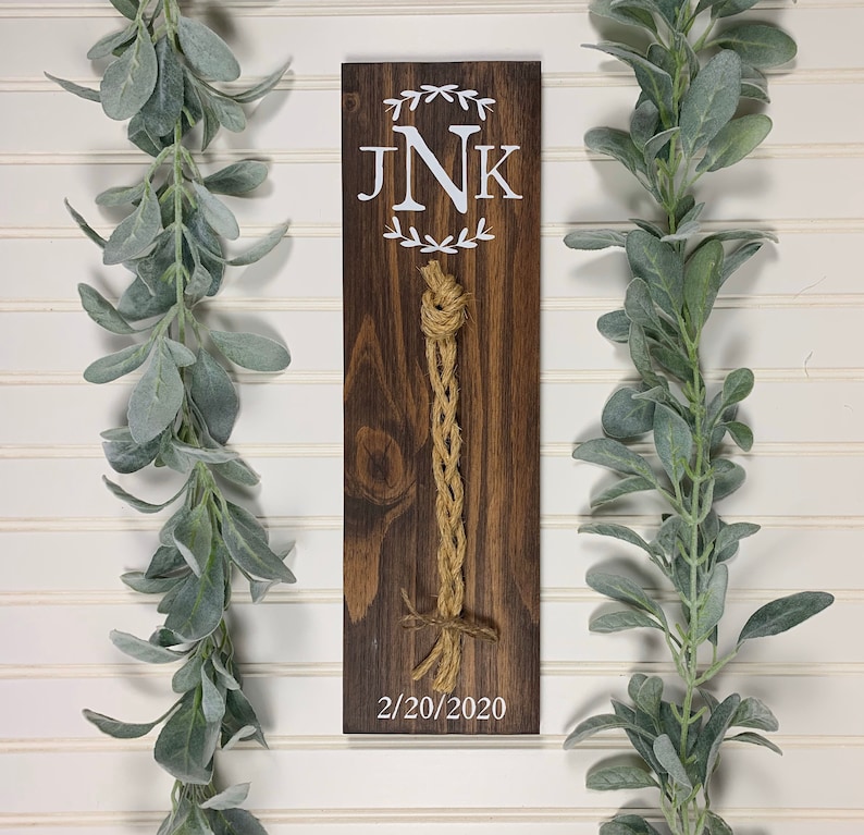 May include: A wooden sign with a brown stain and white lettering. The sign has a monogram of the letters "JNK" with a wreath of leaves around the letters. The date "2/20/2020" is written below the monogram. A brown and beige rope is attached to the sign.