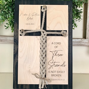 May include: A wooden sign with a black frame and a light wood background. A rope is woven into a cross shape with the text "A cord of three strands is not easily broken. Ecclesiastes 4:12" and the names "Nakia & Rohan Black" and "July 6, 2023" written on the sign.