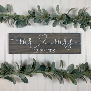 May include: A dark grey wooden sign with the words "mr & mrs" in white script, joined by a heart. The date "12.29.2018" is below. The sign is set against a white background, decorated with green leafy garlands.
