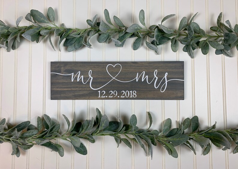 May include: A dark grey wooden sign with the words "mr & mrs" in white script, joined by a heart. The date "12.29.2018" is below. The sign is set against a white background, decorated with green leafy garlands.