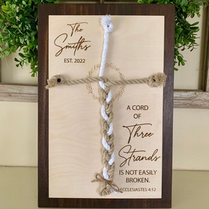 May include: A wooden plaque with a brown frame and a light wood background. The plaque features a white and brown braided rope in the shape of a cross. The text "The Smiths EST. 2022" is engraved at the top of the plaque. The text "A cord of three strands is not easily broken. Ecclesiastes 4:12" is engraved below the rope.