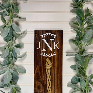 May include: A dark brown wooden sign with the initials "JNK" in white lettering surrounded by a wreath design. A natural twine rope hangs from the bottom of the sign.