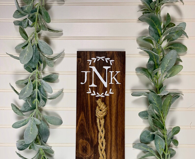 May include: A dark brown wooden sign with the initials "JNK" in white lettering surrounded by a wreath design. A natural twine rope hangs from the bottom of the sign.