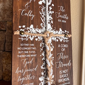 May include: A rectangular dark brown wooden sign featuring a white floral cross design and three braided ropes in white and tan. The text reads Jordyn & Colby, The Smiths EST. 2026, and verses from Matthew 19:6 and Ecclesiastes 4:12 in white lettering.