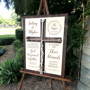 May include: A wooden sign with a brown background and white text. The sign features a braided rope with knots and the text "Sidney & Mykia" and "The Sapps 07/11/24". The sign also includes a quote from the Bible, "So they are no longer two, but one flesh. Therefore what God has joined together, let no one separate. Matthew 19:6" and "A cord of three strands is not easily broken. Ecclesiastes 4:12".