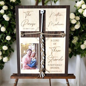 May include: A wooden sign with a cross made of rope, a photo of a couple, and the text "The Piercys Est. May 17, 2025", "Nathan & Madissen", "A Cord of Three Strands Is Not Easily Broken. Ecclesiastes 4:12".