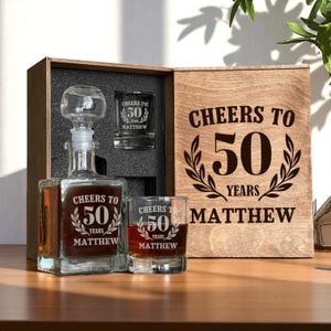 Puede incluir: Un juego de regalo de madera con una jarra, dos vasos de chupito y una caja de madera. La jarra y los vasos de chupito tienen grabado "Cheers to 50 Years Matthew". La tapa de la caja también tiene el mismo texto.