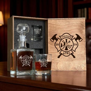 Pode incluir: Um conjunto de presente de madeira com um decantador, dois copos de shot e um copo de whisky. O decantador e os copos são gravados com um emblema de bombeiro. A caixa de madeira tem um emblema correspondente, o nome "Johnson" e os números "20" e "24".