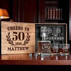 May include: A wooden gift set with a decanter, three glasses, and whiskey stones. The set is engraved with "Cheers to 50 Years Matthew." The decanter and glasses also feature the same inscription. The set is displayed in a wooden box.