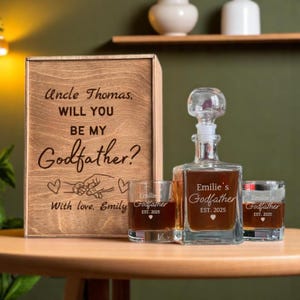 May include: A wooden box with the text "Uncle Thomas, WILL YOU BE MY Godfather?" is next to a glass decanter and two glasses. The decanter and glasses are engraved with "Emilie's Godfather EST. 2025". The box also has the text "With love, Emily".