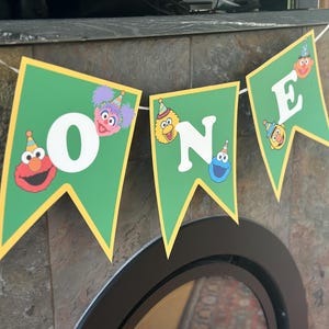May include: A green banner with the word "ONE" spelled out in white letters. Each letter is on a separate pennant, and each pennant has a different Sesame Street character. The pennants are trimmed in yellow and strung together with white string.