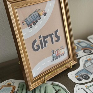 May include: A framed print with a gold-colored frame, featuring a watercolor illustration of a dump truck and a train on a road. The word "GIFTS" is prominently displayed. Additional vehicle cutouts are visible.