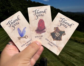 Può includere: Tre biglietti di ringraziamento in stile vintage con le parole "Thank you" in elegante scrittura. Ogni biglietto presenta un design diverso: una chiave con le ali, una gemma rossa e uno stemma con la lettera "H". I biglietti recitano anche "For a magical time!"