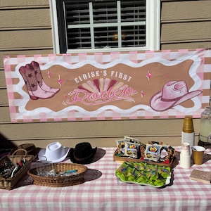 May include: A banner with the text "Eloise's First Rodeo" and illustrations of cowboy boots and a hat. A table is set with hats, snacks, and drinks, covered with a pink and white checkered tablecloth. The scene suggests a rodeo-themed party.