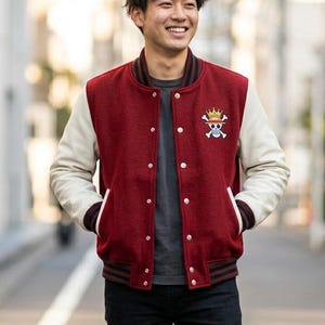 May include: A maroon and cream-colored varsity jacket with a button-up front. The jacket features a skull and crossbones logo with a crown on the chest. The sleeves are cream-colored, and the jacket has a striped collar and cuffs.