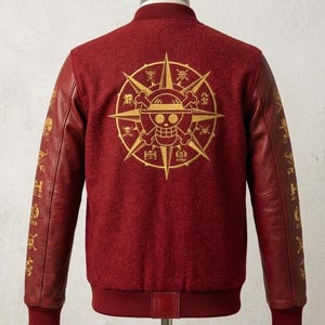 May include: Maroon bomber jacket with gold detailing. The back displays a large gold emblem of a skull and crossbones within a compass design. The sleeves are a darker red leather with gold symbols.