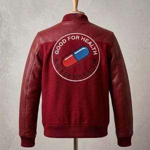 Könnte beinhalten: Burgunderfarbene Bomberjacke mit roten Lederärmeln und geripptem Saum. Auf der Rückseite befindet sich ein kreisförmiger Aufnäher mit einer rot-blauen Pillengrafik und dem Text "GOOD FOR HEALTH, BAD FOR EDUCATION."