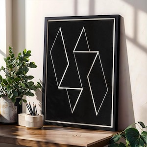 Op de afbeelding: Zwartgelijst kunstwerk met een geometrisch ontwerp van witte lijnen op een zwarte achtergrond. Het kunstwerk staat op een houten oppervlak, naast een plant in een pot en een potloodhouder.