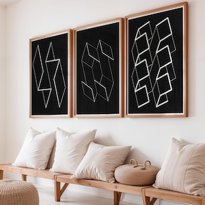 May include: Three framed abstract art prints with geometric white line designs on a black background. The prints are displayed above a wooden bench with beige cushions and a woven basket. The frames are a light brown colour.